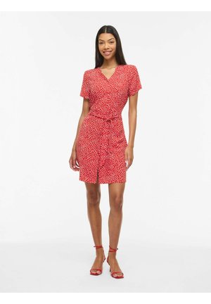 Femme portant une robe portefeuille rouge à manches courtes avec des pois blancs, nouée à la taille, associée à des talons rouges à bride à la cheville, debout sur un fond blanc.