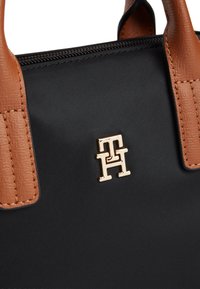 Μαύρη τσάντα χειρός με καφέ δερμάτινες λαβές και χρυσό μεταλλικό μονόγραμμα λογότυπο στο κέντρο κάτω από το φερμουάρ.