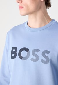 Light blue sweatshirt featuring a large "BOSS" logo. The logo combines solid black and striped gray sections. Classic crew neck design.