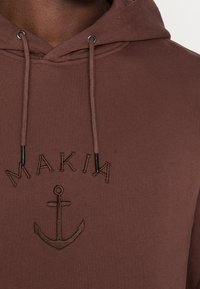 Hnědá mikina s ribbovanou texturou, zdobená vyšitým logem "MAKIA" a symbolem kotvy uprostřed. Obsahuje šňůrky s černými hroty.