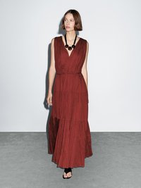 Long, rust-colored maxi dress with a deep V-neck, pleated texture, and a gathered waist. Paired with a black beaded necklace and strappy sandals.