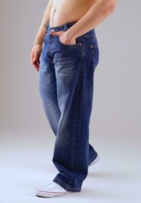 Hemdlose Person in lockeren dunkelblauen Jeans mit einer Hand in der Tasche und weißen Turnschuhen vor einem hellen Hintergrund.