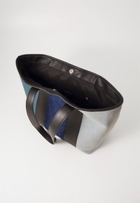 Džínová taška přes rameno s černými koženými uchy a lemem, obsahující modré a šedé panely. Interiér je vyložen černou tkaninou a má zapínání na patentku.