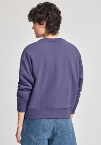 Lila bomullströja med en avslappnad passform, ribbade muddar och fåll. Har en rund halsringning och en mjuk yta. Bärs tillsammans med ljusblå jeans.