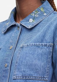 Veste en denim avec boutons sur le devant, grande poche poitrine et col brodé de motifs floraux blancs et verts.