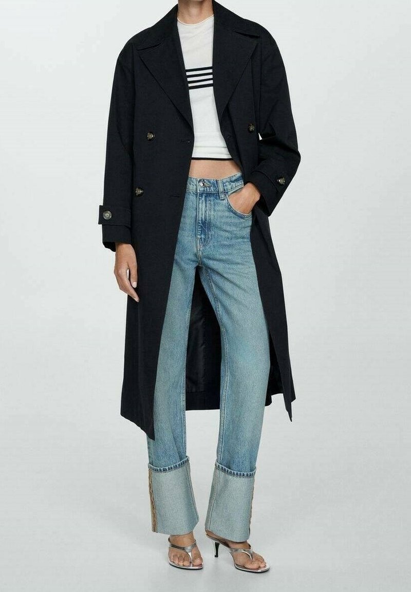 Trench-coat noir croisé porté sur un haut court blanc à rayures noires, associé à un jean évasé bleu clair avec des ourlets bruts. Sandales argentées.