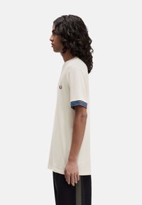 Camisa de color crema con puños a rayas azul y negro, que presenta un pequeño logo bordado en el pecho. Textura suave, ajuste casual.