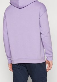 Sudadera morada con una capucha amplia, puños y dobladillo acanalados. Tela suave y texturada; corte relajado, mostrada desde la parte posterior.
