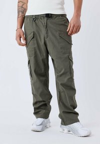 Pantaloni cargo verde oliva con più tasche laterali, fascia elastica in vita e cintura nera. Indossati con sneakers bianche per un look casual.