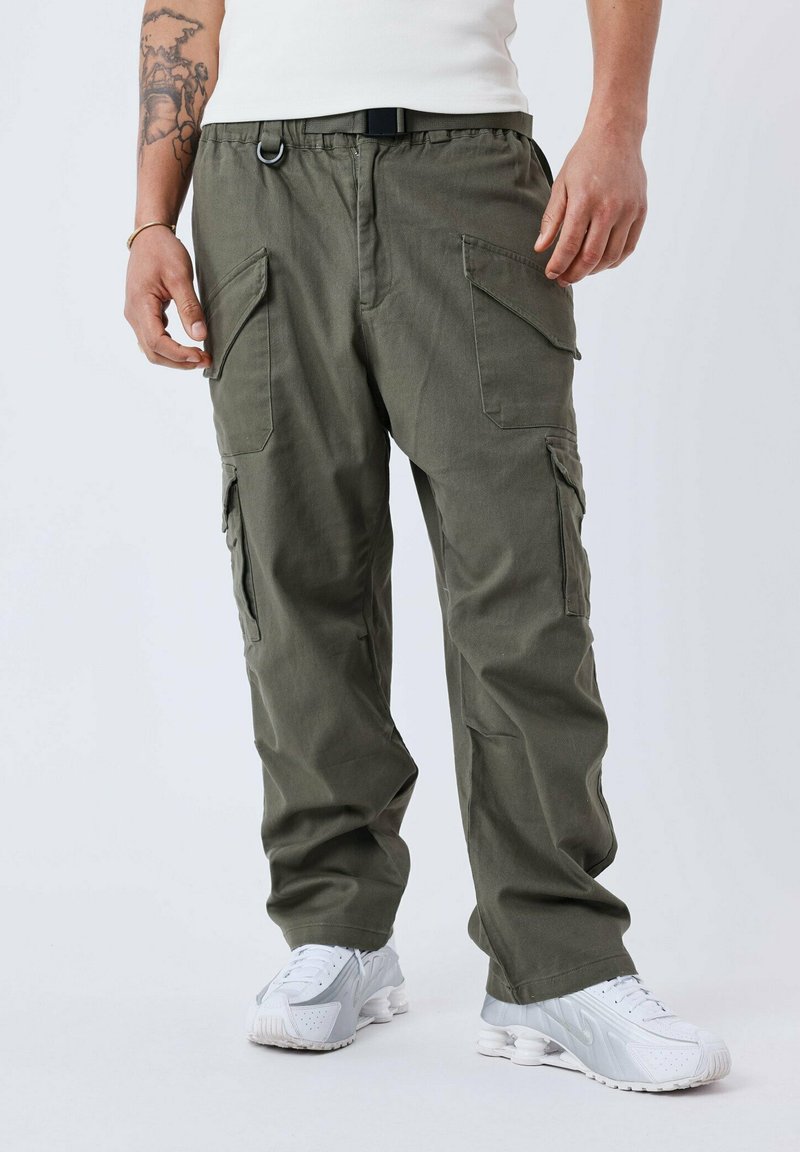 Pantaloni cargo verde oliva con più tasche laterali, fascia elastica in vita e cintura nera. Indossati con sneakers bianche per un look casual.