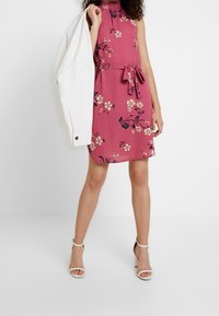 Žena v bezrukávových růžových květinových šatech s páskem v pase, bílé sandály na vysokém podpatku, drží bílou bundu přes jedno rameno.