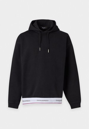 Giovane uomo in piedi che indossa una felpa con cappuccio nera con rifiniture bianche del logo e pantaloni neri larghi con scarpe rosso scuro su sfondo bianco.