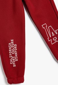 Rote Sweatpants aus weichem Material mit weißem grafischem Text: „CALIFORNIA SPORT CULTURE STANFORD“ und einem großen „L.A.“-Design.