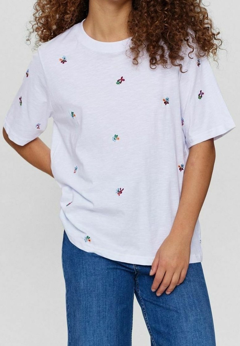 Personne aux cheveux bouclés portant un t-shirt blanc à manches courtes avec de petites fleurs brodées colorées et un jean bleu en denim.
