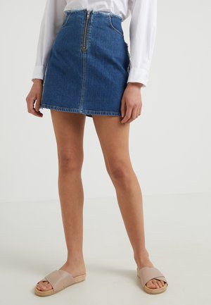 Femme portant une mini-jupe en denim bleu avec fermeture éclair devant, chemise blanche à manches longues et sandales beige, debout sur un fond uni.
