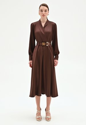 Donna in piedi che indossa un vestito midi marrone a maniche lunghe con cintura in vita e sandali dorati con tacco alto, su sfondo bianco semplice.