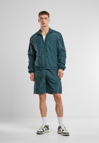 Teal treningsjakke og shorts med hvite piping-detaljer. Lett stoff, glidelås foran, elastisk midjebånd, kombinert med hvite sokker og flerfargede joggesko.