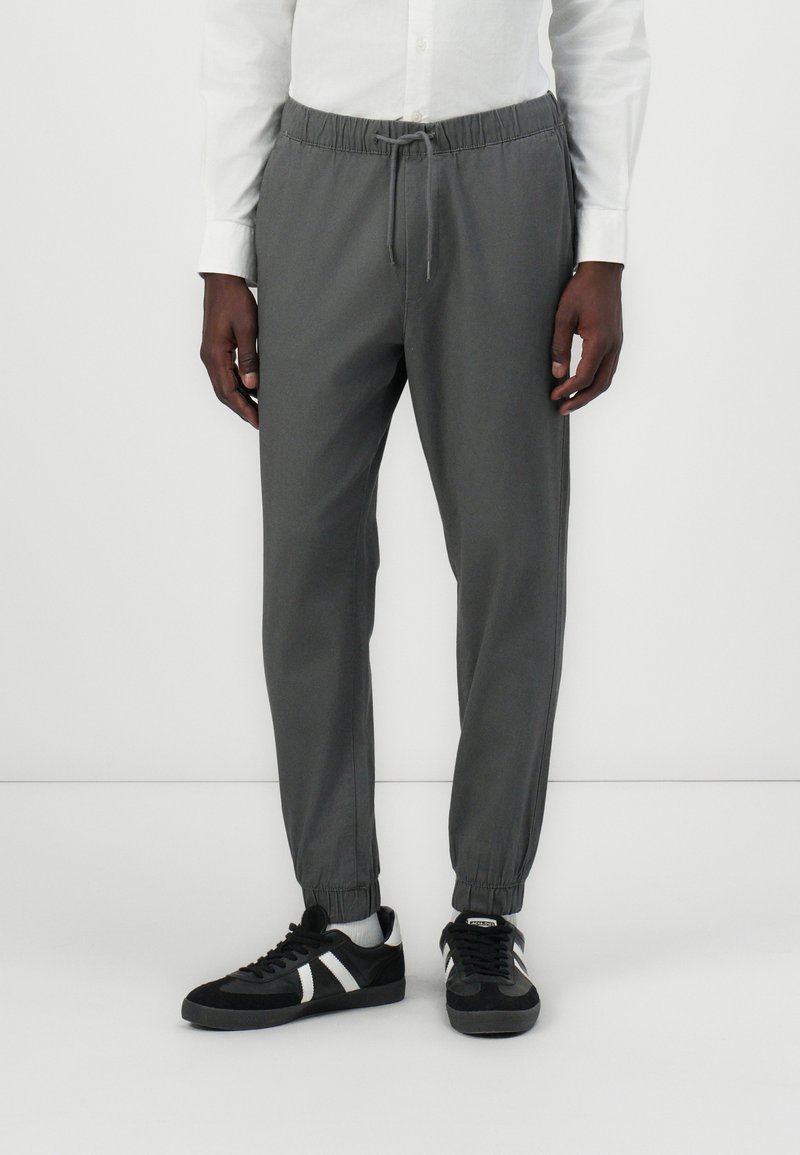 Gray tapered joggers made of soft fabric, featuring an elastic waistband, adjustable drawstring, and elastic cuffs, paired with black sneakers.