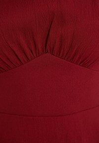 Röd, texturerad klädfiber med en samlad byst och en åtsittande midjeband. Slät yta med subtila linjer och kurvor.