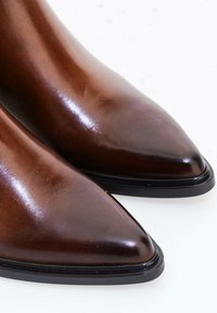 Paire de chaussures en cuir marron poli à bout pointu avec semelles noires sur une surface blanche.