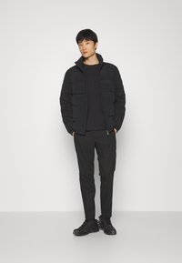 Jachetă puffer neagră cu guler înalt, închidere cu fermoar, purtată peste un top negru și asociată cu pantaloni și pantofi negri, creând un look monocrom.
