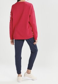 Sudadera roja con hombros caídos y texto blanco en la manga, combinada con pantalones jogger azul marino y zapatillas ligeras de color beige.