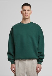 Felpa verde a girocollo in un tessuto strutturato con spalle scese e maniche larghe, abbinata a pantaloni sartoriali chiari.