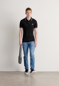 Polo noir avec accents blancs, jeans bleu clair, baskets noires et sac à épaule gris, se tenant devant un fond blanc uni.