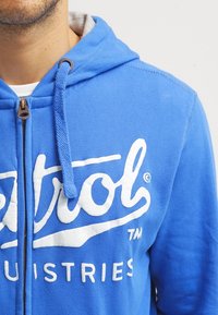Sudadera con cremallera azul hecha de algodón, con un forro interior gris, gran logo blanco en el frente y cordones en la capucha.