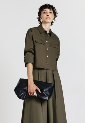 Femme aux cheveux courts et bouclés, portant une chemise à manches longues vert olive et une jupe, tenant un grand sac à main noir avec des anneaux en argent aux doigts.