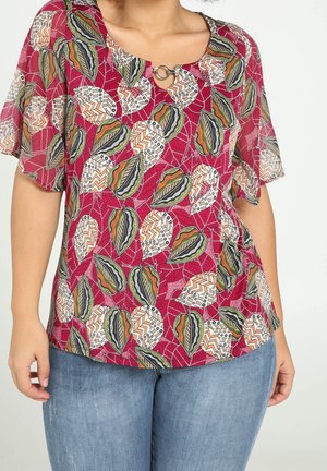 Kobieta ubrana w czerwoną bluzkę z krótkim rękawem, ozdobioną wielokolorowym, abstrakcyjnym wzorem liści oraz srebrnym zapięciem przy dekolcie, w połączeniu z niebieskimi dżinsami.