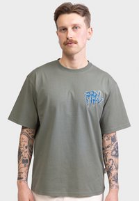 Camiseta de manga corta color verde oliva, hecha de algodón, con un gráfico de gotas azules en el área del pecho izquierdo. Ajuste regular y cuello redondo.