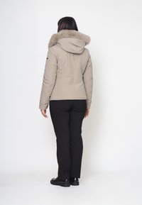 Giacca puffer beige con cappuccio foderato in pelliccia, texture liscia e design leggermente aderente. Abbinata a pantaloni e scarpe neri, vista da dietro.