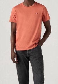 T-shirt corail à manches courtes en coton texturé, avec un col rond et un petit logo rouge Levi's sur la poitrine gauche. Porté avec un jean foncé.