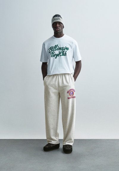 Λευκό πουκάμισο με κοντά μανίκια και πράσινο κείμενο «Billionaire Boys Club», μπεζ παντελόνι φόρμας με κόκκινο γραφικό και μαύρες παντόφλες με σχέδιο.