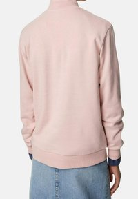 Jasnoróżowa bluza z wysokim kołnierzem, ściągaczami na rękawach i u dołu, wykonana z miękkiego materiału, noszona na spódnicę dżinsową. Prosty, nowoczesny design.