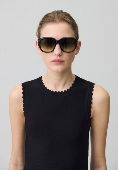 Haut sans manches noir avec un col festonné, porté avec de grandes lunettes de soleil noires à verres dégradés foncés et à monture épaisse.