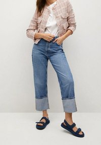 Gehäkelte, pink-weiße kurze Jacke, weißes T-Shirt, hellblaue Jeans mit weitem Bein und kontrastierendem Saum, marineblaue Sandalen mit Riemchen.