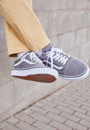 Jambes portant un pantalon beige et des chaussettes blanches avec des baskets grises et blanches, étendues sur un trottoir pavé et un mur en pierre en arrière-plan.