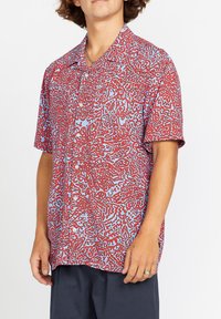 Camisa de botones de manga corta en azul claro con un patrón abstracto rojo, con cuello tipo campamento y corte relajado. Textura de tela suave.