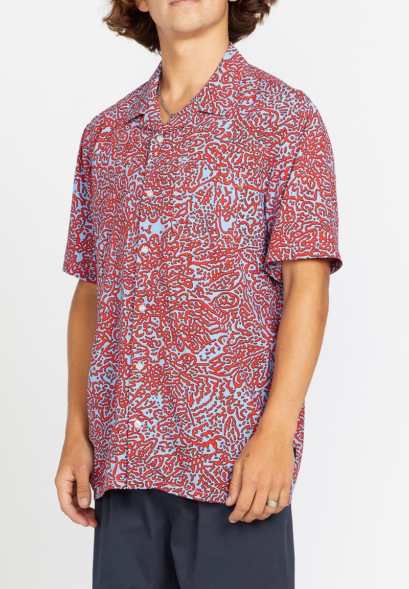 Camisa de botones de manga corta en azul claro con un patrón abstracto rojo, con cuello tipo campamento y corte relajado. Textura de tela suave.