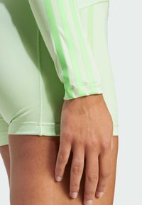 Maglietta sportiva verde menta con tre strisce verticali, texture morbida, design aderente e dettagli a costine sui polsini. Ingrandimento del braccio e del tessuto.