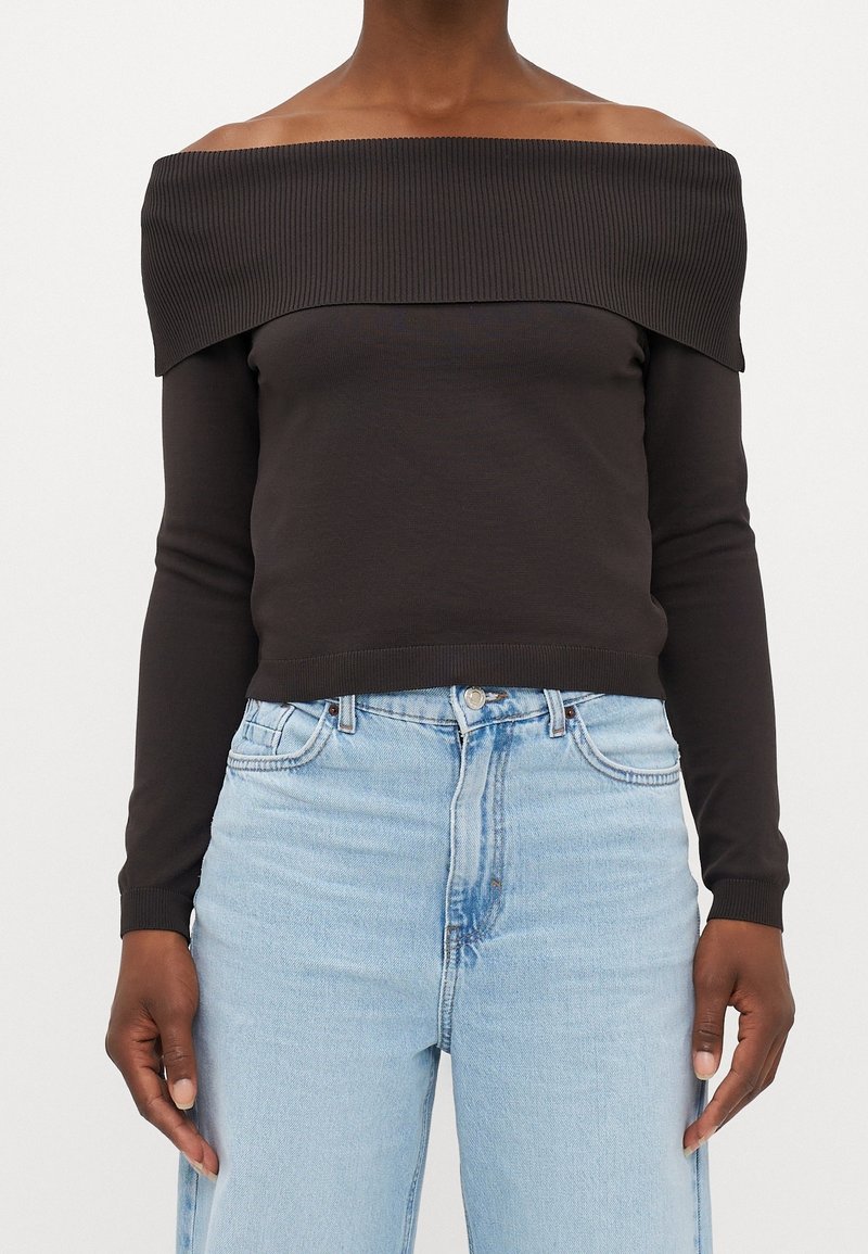 Pull marron foncé à épaules dénudées avec détails côtelés ; manches longues, coupe ajustée, associé à un jean taille haute bleu clair.