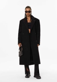 Manteau noir croisé avec larges revers, porté sur un haut court, associé à un pantalon ample avec des fentes sur les côtés et des baskets noires épaisses.