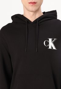 Felpa nera con cappuccio realizzata in tessuto morbido, con tasca frontale, cappuccio regolabile e logo CK bianco sull'area sinistra del petto.