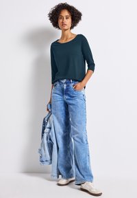 Mörk teal långärmad topp, lös passform, i kombination med ljusblå vida jeans. Modell som håller en ljusare blå denimjacka, iförd vita sneakers.