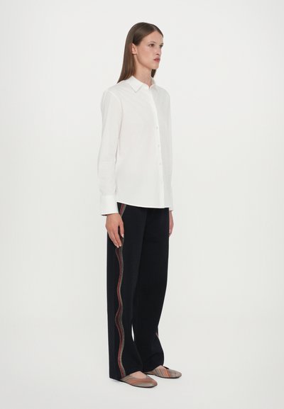 Camicia bianca con maniche lunghe abbinata a pantaloni scuri con una striscia multicolore ondulata. Indossata con scarpe basse a righe.