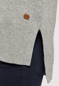 Jasnoszary sweter dzianinowy z żeberkowym dołem, bocznymi rozcięciami i małą brązową skórzaną metką naszytą na dolnym brzegu. Nosi się go na ciemnych spodniach.