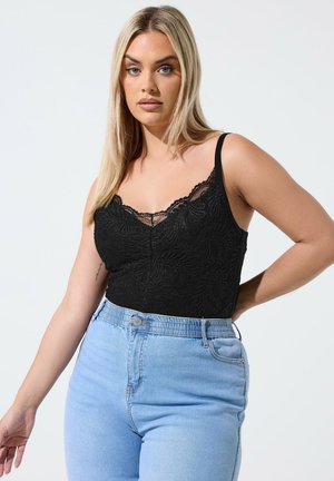 Blonde vrouw met zwart kanten hemdje en lichtblauwe hooggetailleerde spijkerbroek, staande tegen een lichtgrijze achtergrond.