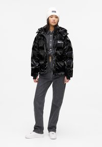 Junge Person mit einer weißen Mütze, glänzender schwarzer Pufferjacke, gebundenem grauen Hemd, dunklen Jeans und weißen Sneakers, die vor einem einfarbigen Hintergrund steht.